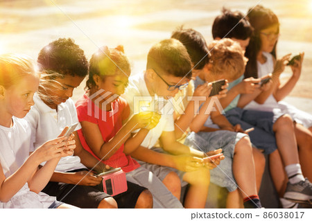 Smiling children sitting at urban street with mobile devices 86038017