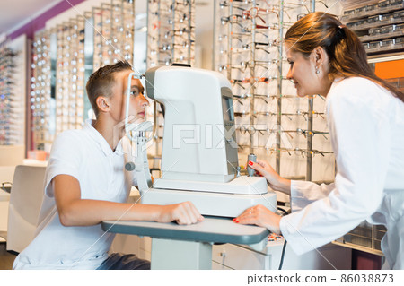 Teenage boy receiving eye examination on modern refractometer in optical store with professional female ophthalmologist 86038873