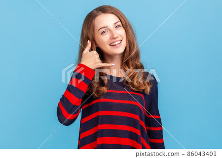 Hey handsome, call me. Smiling woman wearing striped casual style sweater flirting holding fingers near ear showing call gesture, answering call. Indoor studio shot isolated on blue background. 86043401