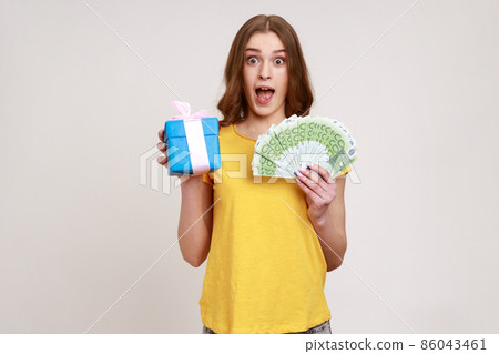 Wow, crazy gift shopping. Surprised girl in yellow casual T-shirt holding present box, euro money banknotes and looking amazed, shocked by purchase. Indoor studio shot isolated on gray background. 86043461