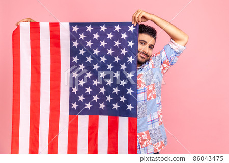Portrait of attractive man holding USA flag in hands, looking at camera with toothy smile, proud of his social security in country, feeling in safe. Indoor studio shot isolated on pink background. 86043475