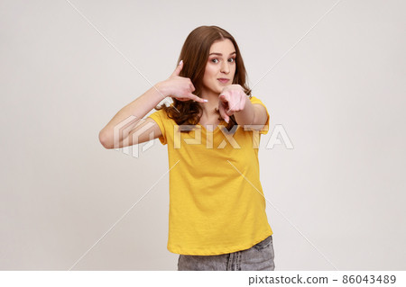 Portrait of beautiful brown haired teenager girl in casual yellow T-shirt standing with call gesture, looking and pointing at camera. Indoor studio shot isolated on gray background. 86043489