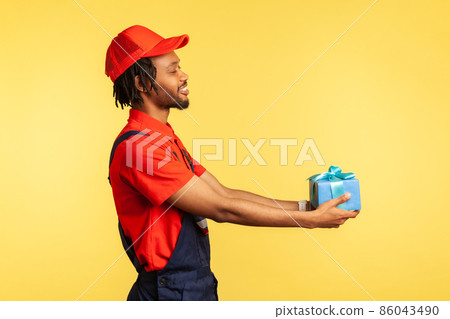 Side view of happy courier in blue overalls and red T-shirt holding wrapped box, smiling friendly to client, carrying birthday present. Indoor studio shot isolated on yellow background. Side view of happy courier in blue overalls and red T-shirt holding wrapped box, smiling friendly to client, carrying birthday present. Indoor studio shot isolated on yellow background. 86043490