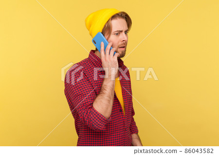 Portrait of hipster bearded guy in beanie hat and checkered shirt talking on phone, having serious mobile conversation, making important call. Indoor studio shot isolated on yellow background. 86043582