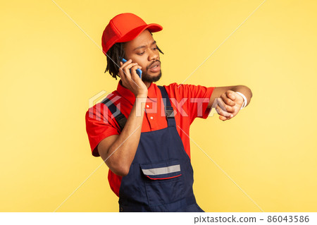 Portrait of concentrated courier wearing blue overalls talking phone with client, looking at smart watch, delivery service on time. Indoor studio shot isolated on yellow background. Portrait of concentrated courier wearing blue overalls talking phone with client, looking at smart watch, delivery service on time. Indoor studio shot isolated on yellow background. 86043586
