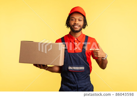 Portrait of cargo transportation, delivery service. Friendly courier in uniform pointing at cardboard box, delivering parcel to client. Indoor studio shot isolated on yellow background. Portrait of cargo transportation, delivery service. Friendly courier in uniform pointing at cardboard box, delivering parcel to client. Indoor studio shot isolated on yellow background. 86043598