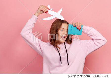Portrait of happy curly haired teenage girl in hoodie winking, holding passport and paper airplane, looking at camera with toothy smile, travelling. Indoor studio shot isolated on pink background 86043621