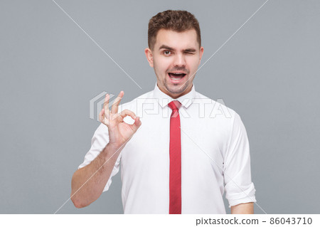 Its Ok. Portrait of young funny handsome man in white shirt and tie standing with Ok sign and looking at camera winking. indoor isolated on gray background. 86043710