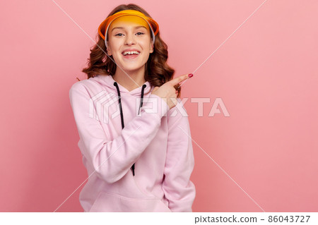Portrait of happy curly haired teenage girl in hoodie and yellow sun visor smiling and pointing at empty space background, place for advertisement. Indoor studio shot isolated on pink background 86043727