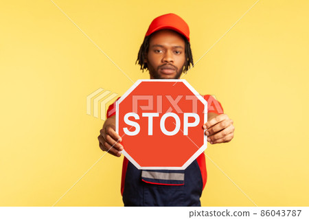 Portrait of serious handyman wearing uniform and red visor cap looking at camera, holding out showing red stop traffic sign, warns of danger. Indoor studio shot isolated on yellow background. 86043787