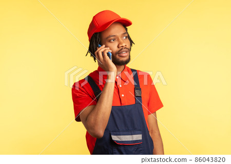 Portrait of courier or repairman in overalls taking phone with client, specify the time and place of completing work or delivery of goods. Indoor studio shot isolated on yellow background. 86043820