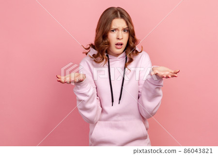 Portrait of unhappy frustrated teen girl with curly brunette hair spreading hands with shock and misunderstanding, how could you sign. Indoor studio shot isolated on pink background 86043821