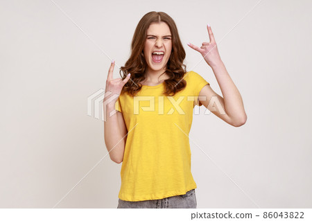Extremely excited satisfied teenager girl with brown hair in yellow T-shirt showing rock and roll gesture with fingers, having fun, resting. Indoor studio shot isolated on gray background. 86043822