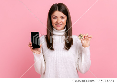 Smiling female holding gold bitcoin and smartphone with empty screen for advertisement or promotion, wearing white casual style sweater. Indoor studio shot isolated on pink background. 86043823