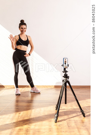 Slim female recording tutorial video for fitness vlog, waving hand, saying hi to followers, wearing black sports top and tights. Full length studio shot illuminated by sunlight from window. 86043825