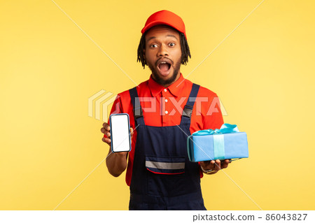 Portrait of surprised courier in blue uniform holding and showing gift boxes and smartphone with empty display, order application. Indoor studio shot isolated on yellow background. Portrait of surprised courier in blue uniform holding and showing gift boxes and smartphone with empty display, order application. Indoor studio shot isolated on yellow background. 86043827