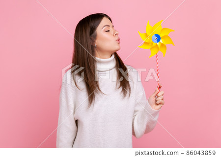 Portrait of carefree brunette woman blowing at paper windmill, playing with pinwheel toy on stick, wearing white casual style sweater. Indoor studio shot isolated on pink background. Portrait of carefree brunette woman blowing at paper windmill, playing with pinwheel toy on stick, wearing white casual style sweater. Indoor studio shot isolated on pink background. 86043859