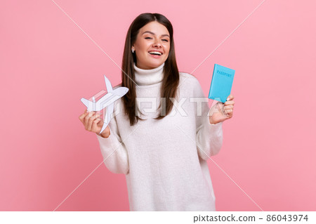 Portrait of excited female with passport and paper plane in hands rejoices future tour, looking at camera., wearing white casual style sweater. Indoor studio shot isolated on pink background. 86043974