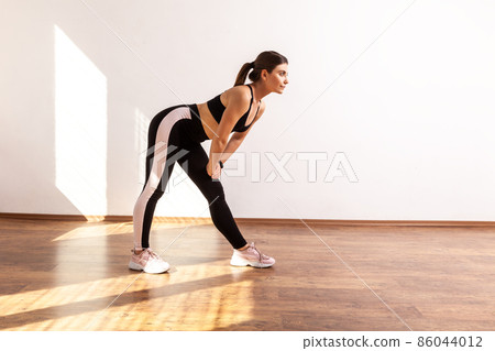 Full length portrait of warming up before training, touching leg with both arms, stretching, wearing black sports top and tights. Indoor studio shot illuminated by sunlight from window. 86044012