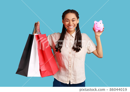 Positive optimistic woman with black dreadlocks holding shopping bags and piggy bank, cashback from buying purchases, wearing white shirt. Indoor studio shot isolated on blue background. 86044019