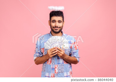Portrait of charming angelic bearded man with halo over head holding fan of dollar bills and smiling happily, happy to have money. Indoor studio shot isolated on pink background. 86044040