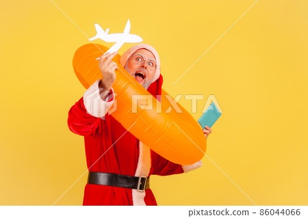 Portrait of amazed elderly man in santa claus costume standing with rubber ring, passport and paper plane, being happy to travel on Christmas holidays. Indoor studio shot isolated on yellow background Portrait of amazed elderly man in santa claus costume standing with rubber ring, passport and paper plane, being happy to travel on Christmas holidays. Indoor studio shot isolated on yellow background 86044066