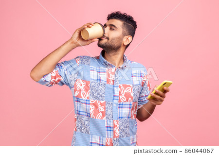Portrait of attractive young adult man in casual style shirt standing and holding disposable cup and phone , drinking and enjoying hot beverage. Indoor studio shot isolated on pink background. 86044067