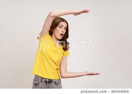 Excited brown haired teen girl showing big empty space, holding box copy space, keeps mouth open, looking at camera with shocked expression. Indoor studio shot isolated on gray background. 86044068