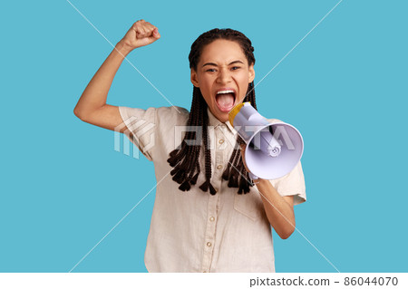 Portrait of woman with black dreadlocks standing with raised hands and holding megaphone, screaming in loud speaker, protesting, wearing white shirt. Indoor studio shot isolated on blue background. 86044070