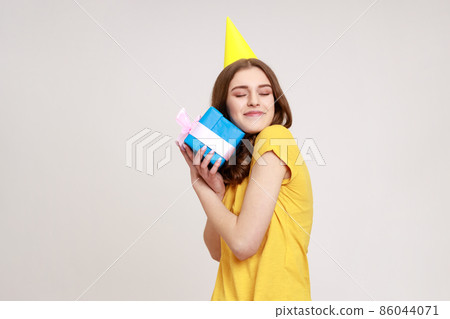 Portrait of happy satisfied teenager girl embracing blue gift box ,receiving surprise, being happy, keeps eyes closed, wearing yellow T-shirt. Indoor studio shot isolated on gray background. 86044071