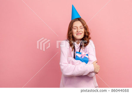 Portrait of joyful smiling curly haired teenage girl in hoodie with party hat hugging wrapped present box, wonder gift, winner. Indoor studio shot isolated on pink background Portrait of joyful smiling curly haired teenage girl in hoodie with party hat hugging wrapped present box, wonder gift, winner. Indoor studio shot isolated on pink background 86044088