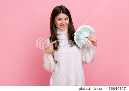 Smiling rich female pointing at big fan of euro banknotes with finger, rejoices of winning lottery, wearing white casual style sweater. Indoor studio shot isolated on pink background. Smiling rich female pointing at big fan of euro banknotes with finger, rejoices of winning lottery, wearing white casual style sweater. Indoor studio shot isolated on pink background. 86044117