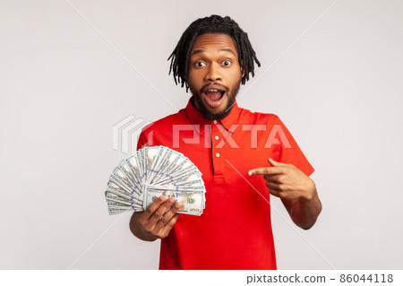 Man with dreadlocks in red casual style T-shirt, excited and surprised to have money, pointing at dollar bills in hand, shocked by big lottery win. Indoor studio shot isolated on gray background. Man with dreadlocks in red casual style T-shirt, excited and surprised to have money, pointing at dollar bills in hand, shocked by big lottery win. Indoor studio shot isolated on gray background. 86044118