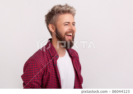 Portrait of joyful caucasian young man with beard and checkered shirt laughing, good mood and humor. Indoor studio shot isolated on gray background 86044123
