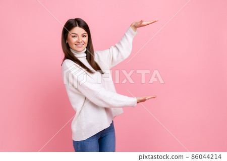 Happy charming woman presenting copy space between hands, holding empty place for commercial text, wearing white casual style sweater. Indoor studio shot isolated on pink background. 86044214
