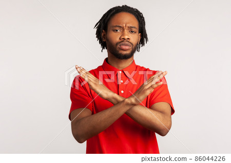 Portrait of young adult bearded man with dreadlocks wearing red T-shirt, crossing hands, gesturing warning or prohibition, meaning stop finish. Indoor studio shot isolated on gray background. 86044226