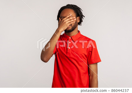 Man with dreadlocks wearing red casual style T-shirt, closing eyes with hand, dont want to see that, ignoring problems, hiding from stressful situations. Indoor studio shot isolated on gray background 86044237