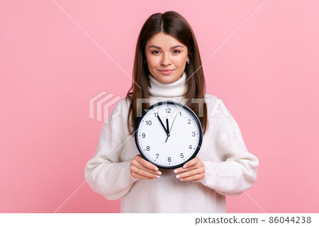 Portrait of serious female holding big wall clock, looking at camera with pleasant smile, time to go, wearing white casual style sweater. Indoor studio shot isolated on pink background. 86044238