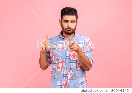 You are loser. Portrait of serious unhappy man in blue casual shirt pointing at camera and showing lame or loser gesture, mocking your failures. Indoor studio shot isolated on pink background. 86044345