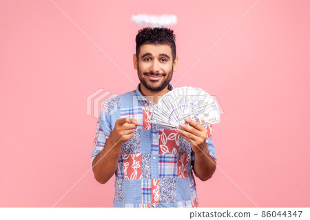 Hey you, make money. Portrait of positive angelic bearded man with halo over head in blue shirt holding fan of dollar bills, pointing finger to camera. Indoor studio shot isolated on pink background. 86044347