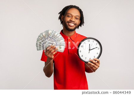 Happy young adult attractive man with dreadlocks wearing red casual style T-shirt, showing dollar cash and wall clock on camera, hourly payment. Indoor studio shot isolated on gray background. Happy young adult attractive man with dreadlocks wearing red casual style T-shirt, showing dollar cash and wall clock on camera, hourly payment. Indoor studio shot isolated on gray background. 86044349