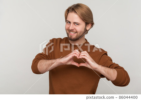 Portrait of happy attractive man with beard wearing sweatshirt, standing with heart or love gesture and looking at camera with toothy smile. Indoor studio shot isolated on gray background. 86044400