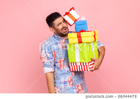 Portrait of funny bearded man carrying lot of heavy present boxes falling on him, looking with frowning face, trying to hold many birthday gifts. Indoor studio shot isolated on pink background. 86044478