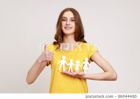 Portrait of happy satisfied female of young age holding paper family chain and showing thumb up, looking at camera with toothy smile. Indoor studio shot isolated on gray background. Portrait of happy satisfied female of young age holding paper family chain and showing thumb up, looking at camera with toothy smile. Indoor studio shot isolated on gray background. 86044480