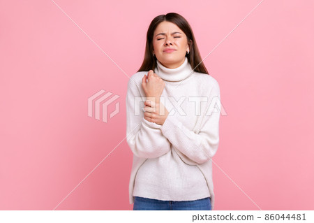 Upset woman standing with grimace of pain, massaging sore wrist, suffering hand injury or sprain, wearing white casual style sweater. Indoor studio shot isolated on pink background. Upset woman standing with grimace of pain, massaging sore wrist, suffering hand injury or sprain, wearing white casual style sweater. Indoor studio shot isolated on pink background. 86044481
