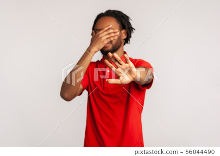 Stop, I don't want to see at this. Young bearded man with dreadlocks wearing red casual style T-shirt, covering his face and showing stop hand gesture. Indoor studio shot isolated on gray background. 86044490