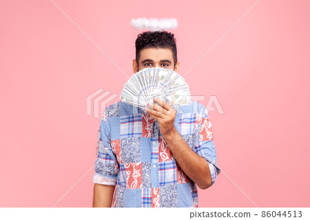 Angelic rich bearded man wearing blue casual style shirt with nimbus on head peeking out of dollar banknotes, rejoicing big profit earn. Indoor studio shot isolated on pink background. 86044513