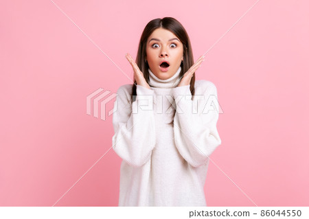 Shocked surprised female with dark hair standing with raised arms, looking at camera with open mouth, wearing white casual style sweater. Indoor studio shot isolated on pink background. 86044550