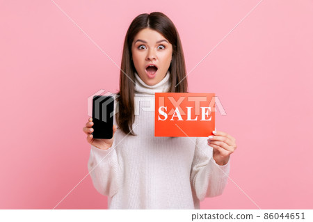Astonished female standing with open mouth, holding sale card and cell phone with empty display, wearing white casual style sweater. Indoor studio shot isolated on pink background. 86044651