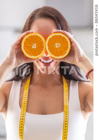 Woman with measuring tape around her neck holds halved orange in front of her eyes 86045916